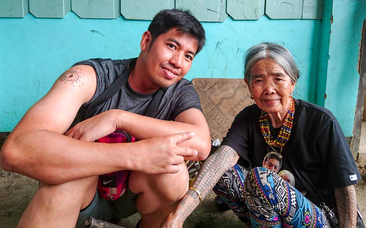Visitor meeting legendary mambabatok Apo Whang-Od in Buscalan, Kalinga. Traditional hand-tapped tattoo artist smiling with traveler in mountain village.